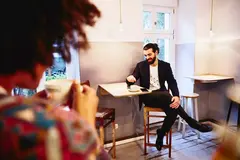 male sitting at a table with coffee, looking down while having a conversation with someone.
