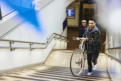 Man carrying a bike up the stairs in a train station. Other man walking up the stairs in the background.