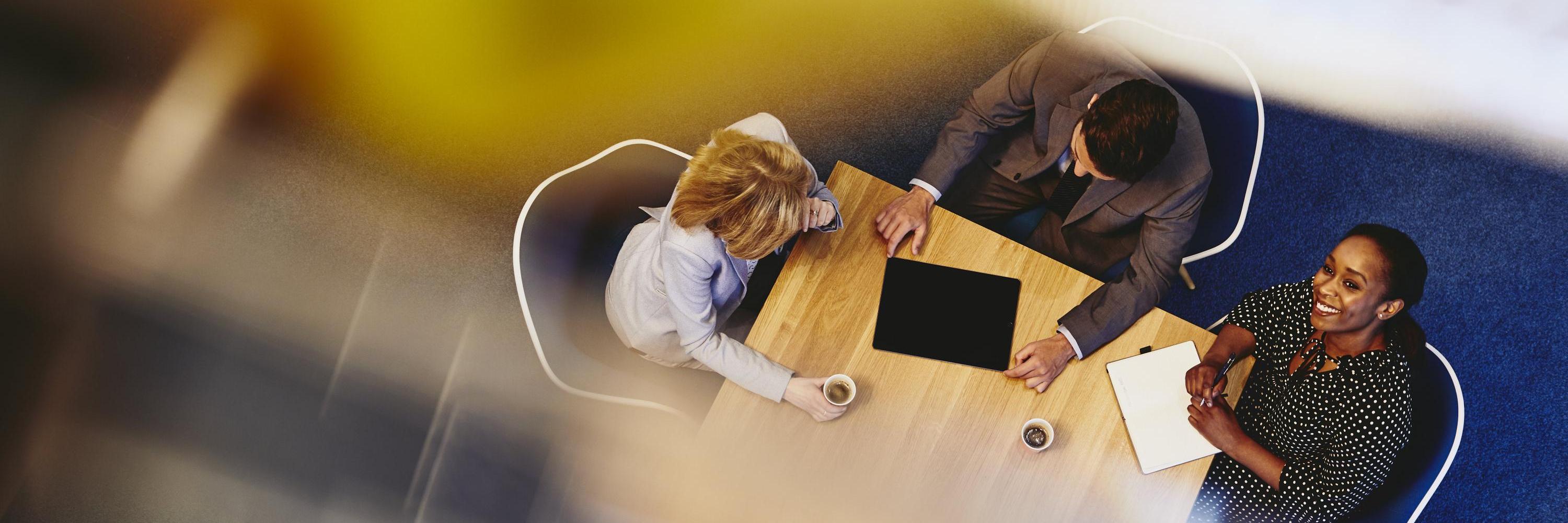 People having a meeting in an office. Woman looking up & smiling. Primary color: yellow. τρεις άνθρωποι συνεδριάζουν
