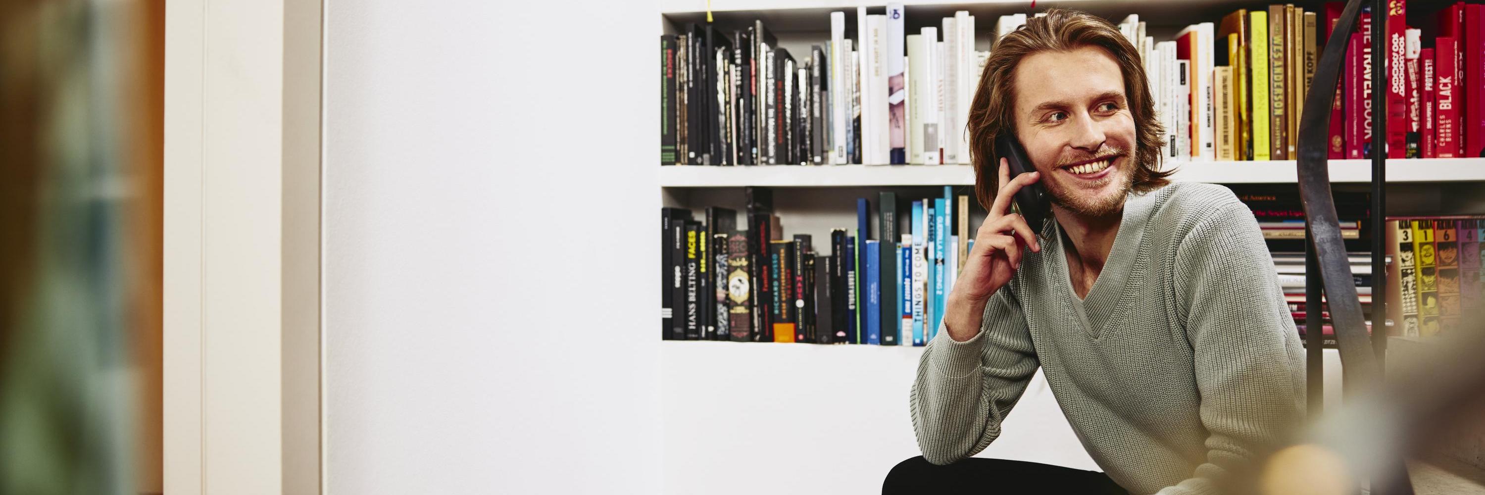 Smiling man sitting on stairs, having a phonecall. bookshelf in background